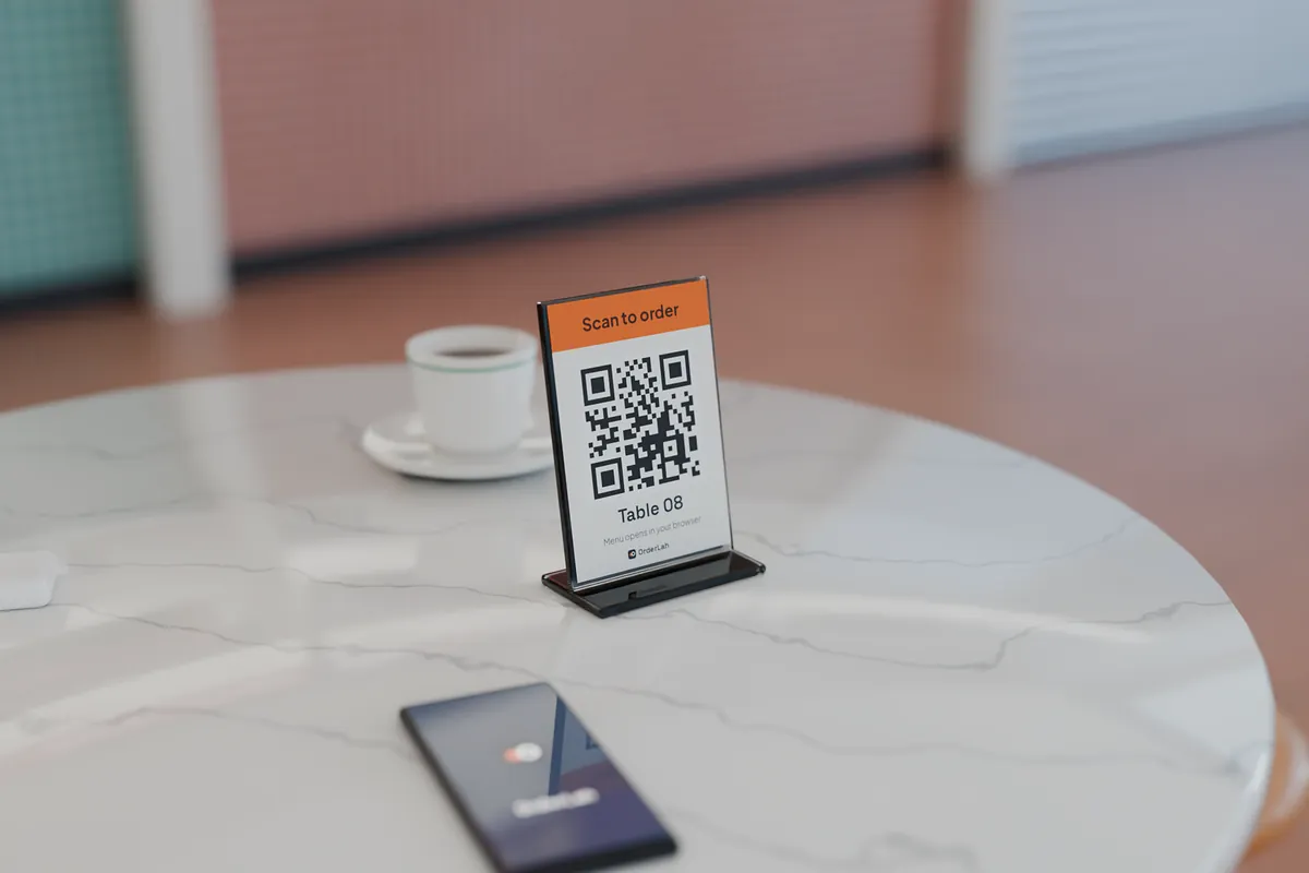 A table card in an acrylic stand on a marble hawker table, printed “Scan to order — Table 08” around a QR code, beside a phone lying face up and a cup of kopi.